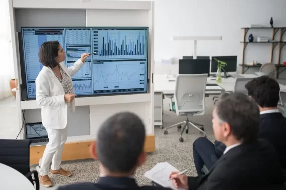 A business woman in a white suit stands in front of a room full of men presenting a business report consisting of various charts and data points.