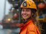 Female engineer. Female offshore engineer wearing a safety helmet and orange protective gear smiles on the deck of an oil rig, with industrial equipment in the background. Female engineer. Female offshore engineer wearing a safety helmet and orange protective gear smiles on the deck of an oil rig, with industrial equipment in the background.